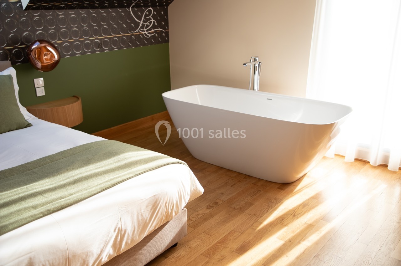 Chambre lumineuse avec baignoire blanche indépendante, parquet en bois et lit avec couverture verte.