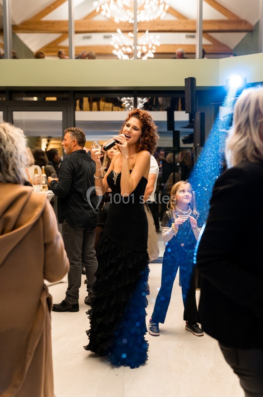 Femme chantant au micro dans une salle éclairée, entourée d'invités debout lors d'un événement festif.