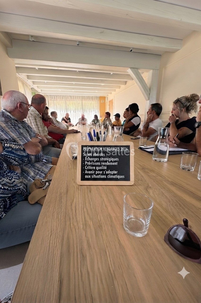 Des personnes assises autour d'une grande table en bois lors d'une réunion, avec un panneau mentionnant des sujets de…