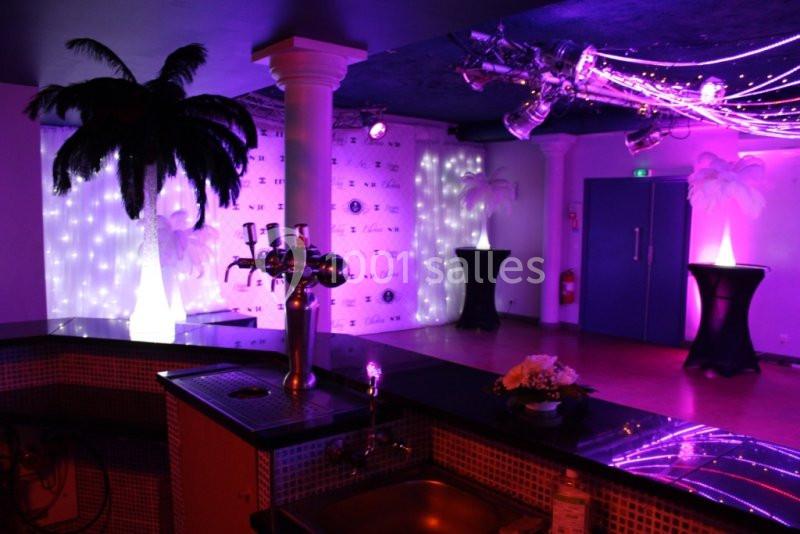 Salle décorée avec des lumières violettes, palmiers artificiels illuminés et comptoir de bar au premier plan.