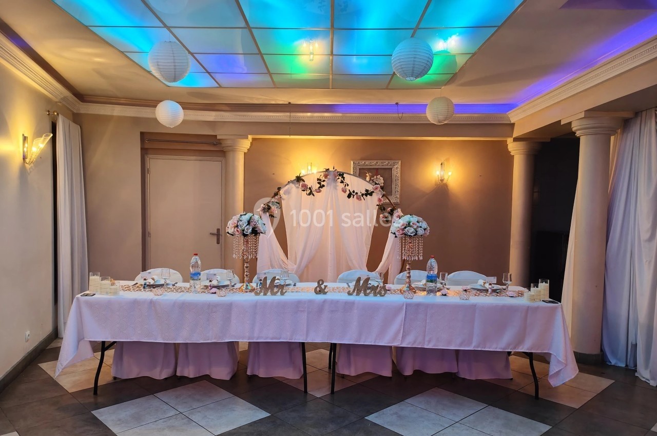 Table de mariage décorée avec nappes blanches, fleurs, arche ornée et éclairage coloré dans une salle de réception.