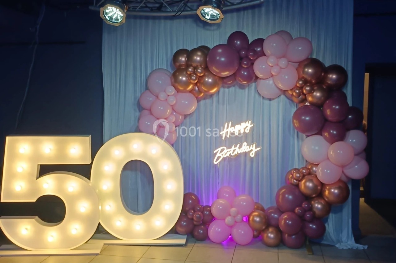 Décoration d'anniversaire avec un ’50’ lumineux, un arc de ballons roses et dorés, et un néon ’Happy Birthday’.