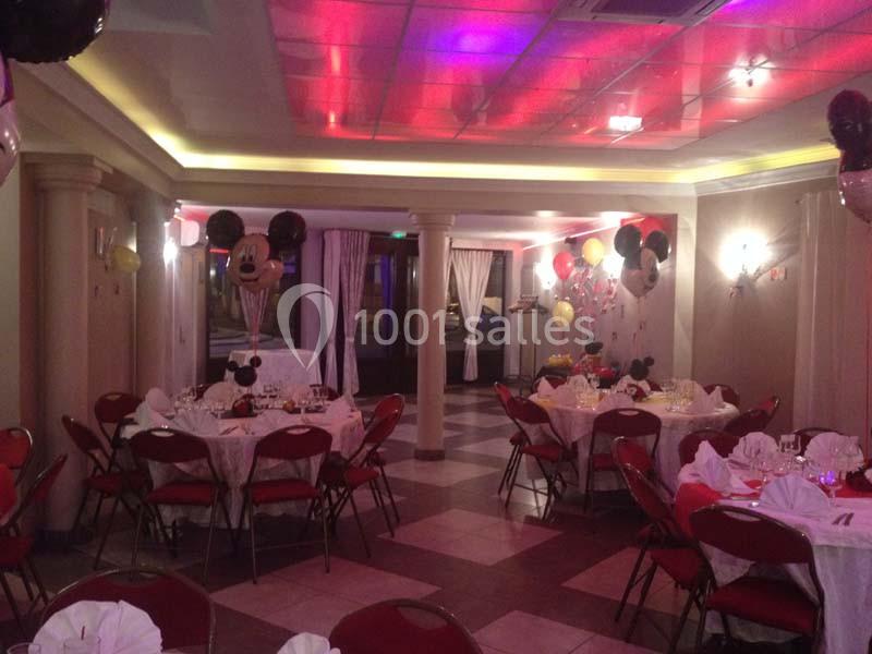 Salle décorée pour une fête avec des ballons Mickey Mouse, des tables dressées et un éclairage coloré au plafond.
