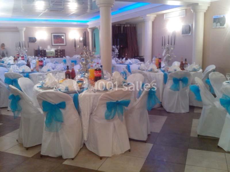 Salle de réception décorée avec des tables rondes, nappes blanches et nœuds bleus sur les chaises.