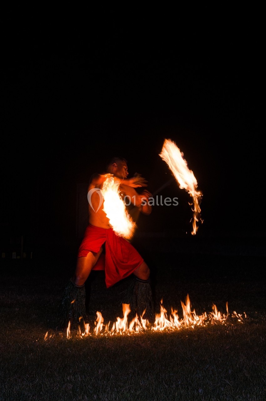 Un homme en pagne rouge jongle avec un bâton enflammé, entouré de flammes au sol, dans l'obscurité.