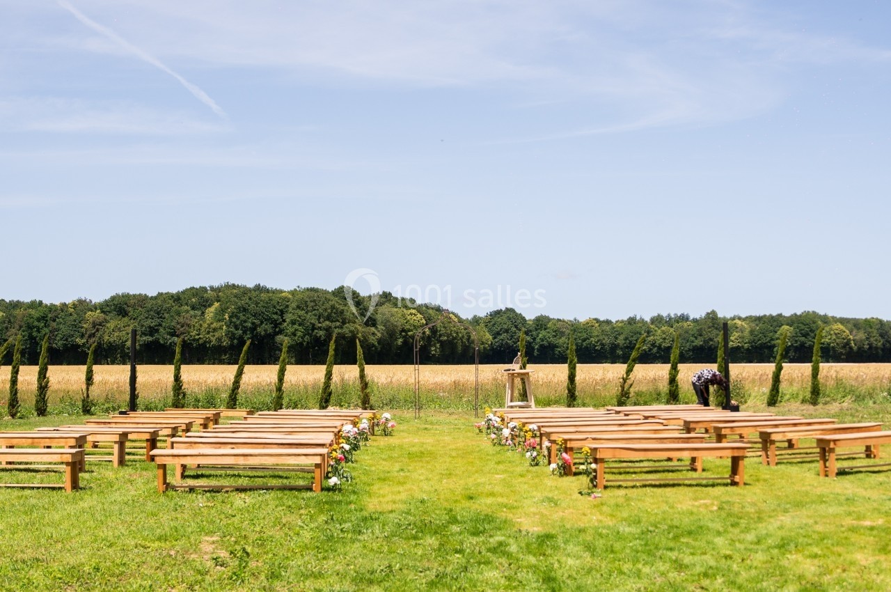 Chaises et bancs en bois disposés en extérieur pour une cérémonie, avec un champ et des arbres en arrière-plan.