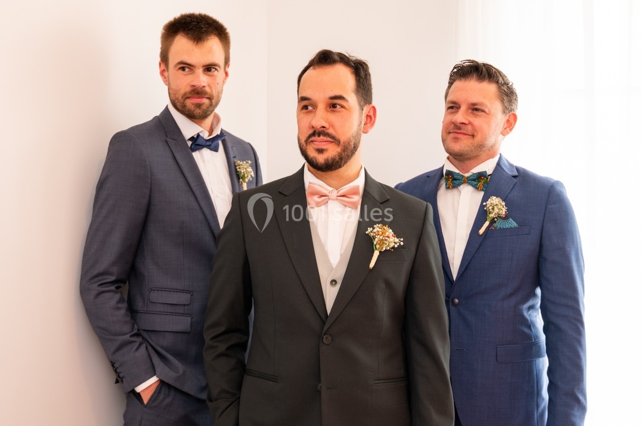 Trois hommes en costume, dont un au centre avec un nœud papillon rose, posent dans un intérieur lumineux.