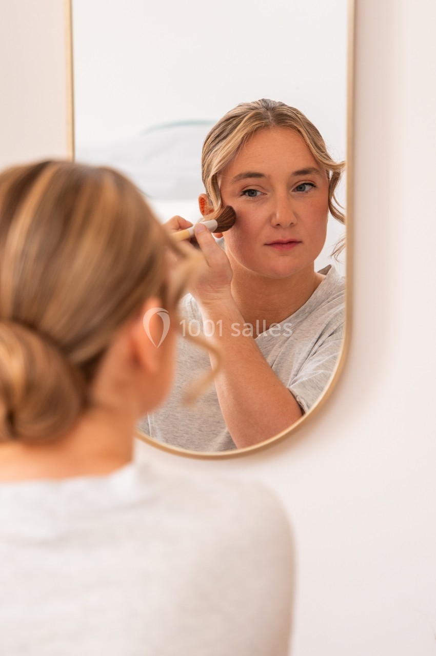 Une femme applique du maquillage sur son visage devant un miroir, dans un environnement lumineux et épuré.
