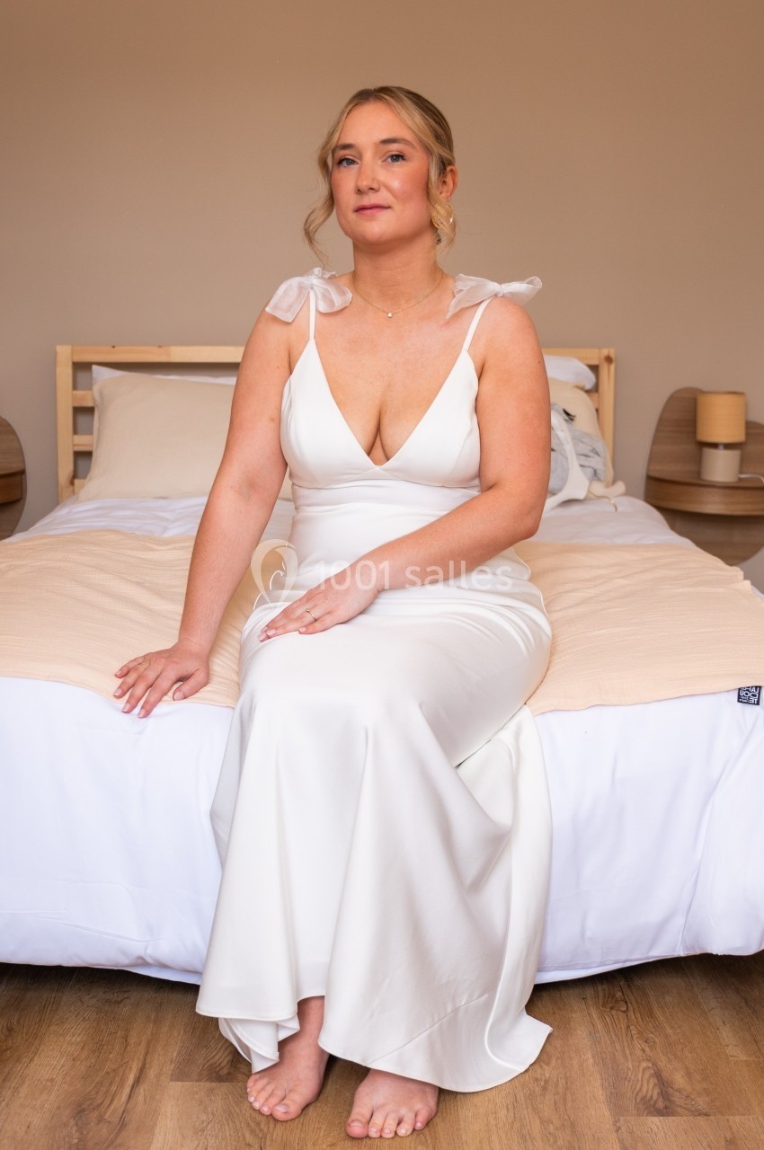 Femme en robe blanche élégante assise sur un lit dans une chambre au décor minimaliste.