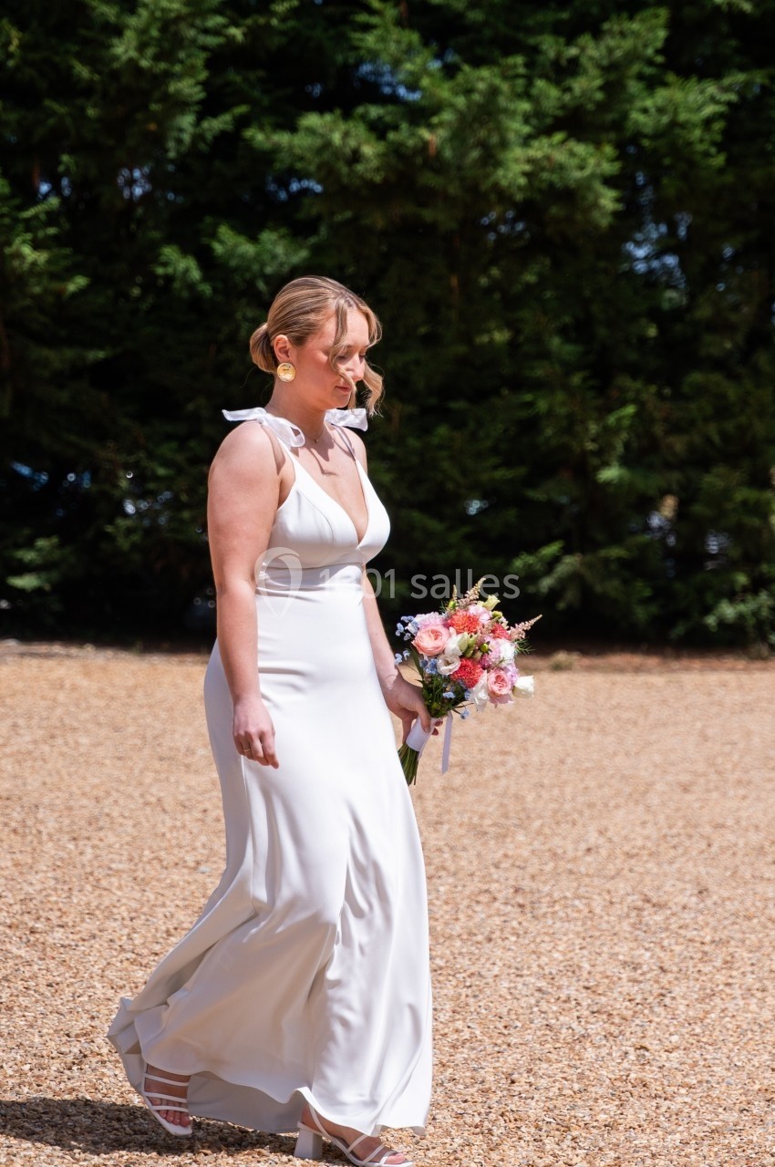 Femme en robe blanche tenant un bouquet de fleurs, marchant sur un chemin de gravier devant des arbres verts.