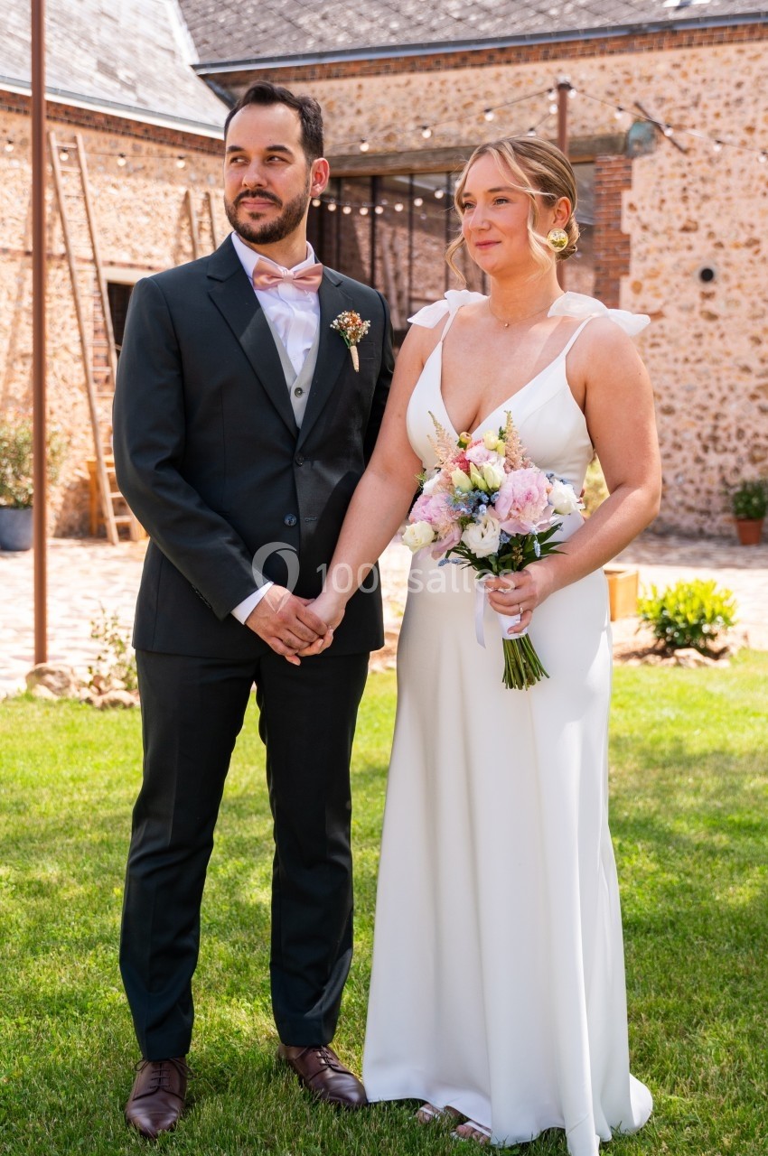 Un couple en tenue de mariage pose dans un jardin ensoleillé devant un bâtiment en pierre.