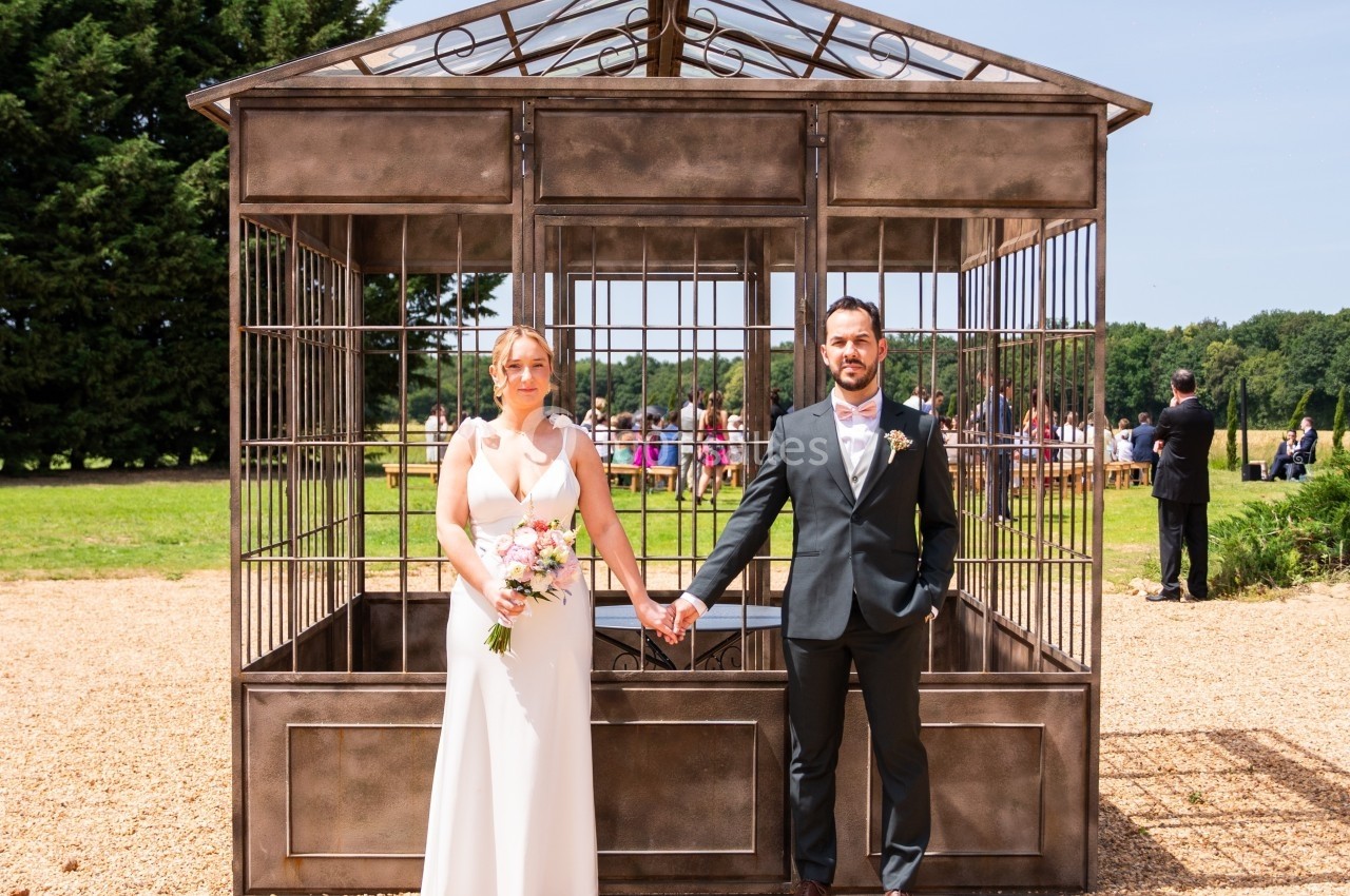 Un couple en tenue de mariage pose devant une structure métallique dans un jardin par temps ensoleillé.
