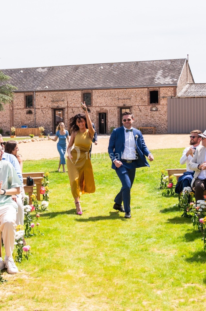 Un couple marche joyeusement dans une allée extérieure bordée de fleurs, entouré d'invités assis.