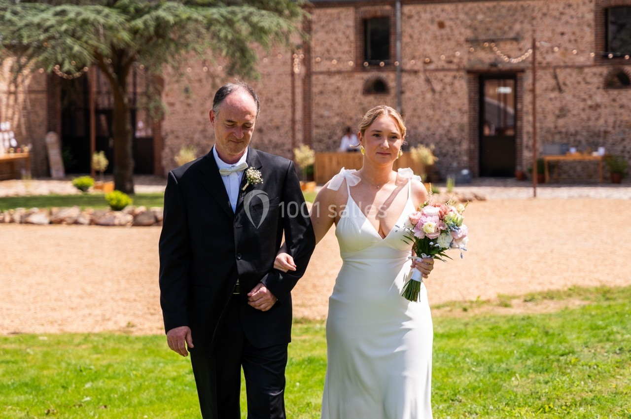 Une mariée en robe blanche marche sur une pelouse, accompagnée d'un homme en costume sombre, devant un bâtiment en pierre.