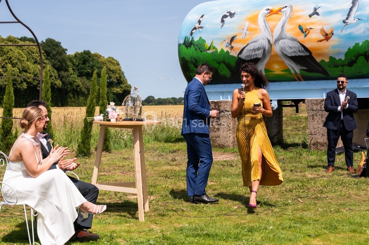 Un couple assis applaudit une femme en robe jaune et un homme en costume lors d'un événement en plein air champêtre.