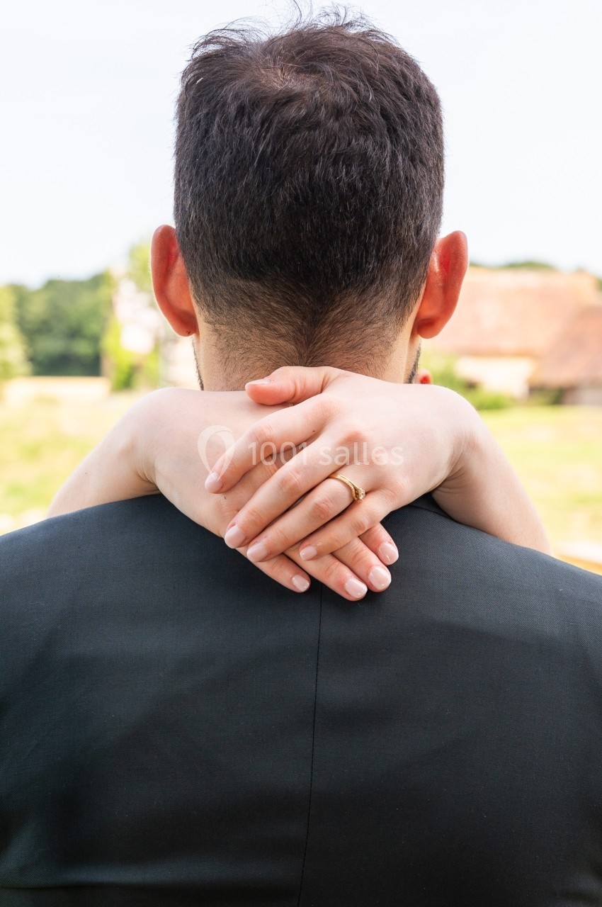 Une femme enlace un homme par le cou, ses mains visibles avec une alliance, en extérieur par temps clair.