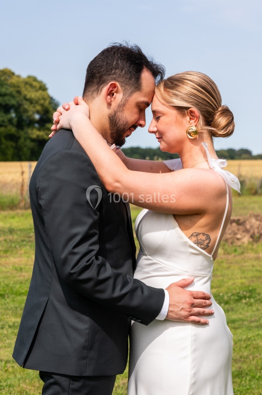 Un couple en tenue de mariage s'enlace tendrement dans un champ sous un ciel dégagé.