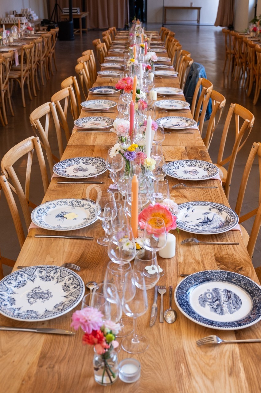 Tables en bois alignées, décorées de vaisselle en porcelaine, fleurs colorées et bougies dans une salle lumineuse.