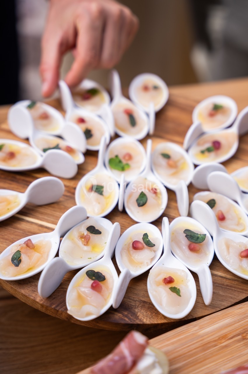 Cuillères en porcelaine contenant des bouchées de carpaccio de poisson, garnies de câpres et de morceaux de fruits.