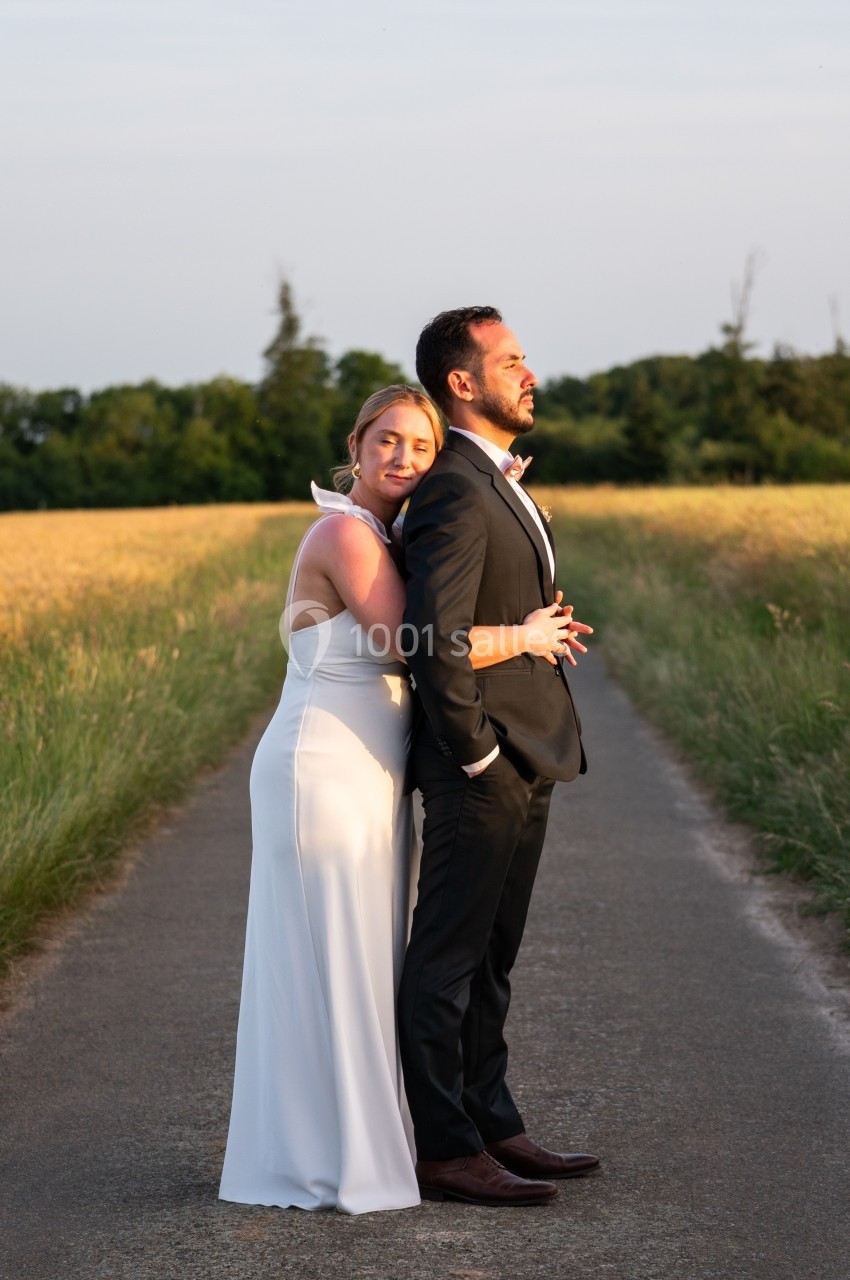 Un couple en tenue de mariage se tient sur un chemin rural, entouré de champs et d'arbres au coucher du soleil.