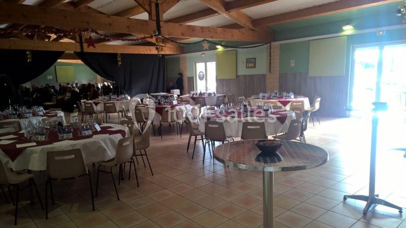 Salle de réception avec tables rondes dressées, nappes blanches et rouges, éclairée par la lumière naturelle.