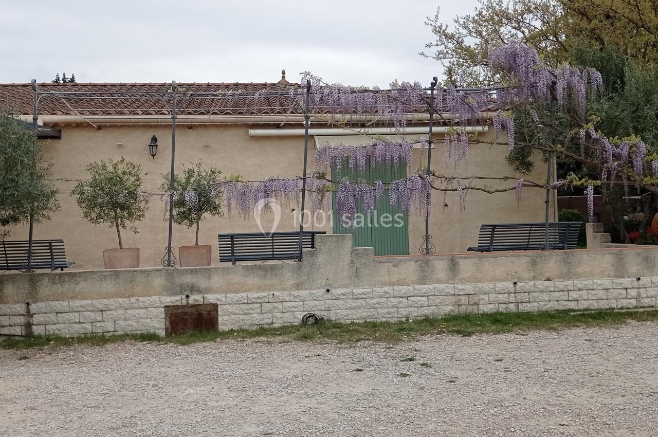Terrasse avec bancs sous une pergola ornée de glycines en fleurs, devant une maison beige avec toit en tuiles.