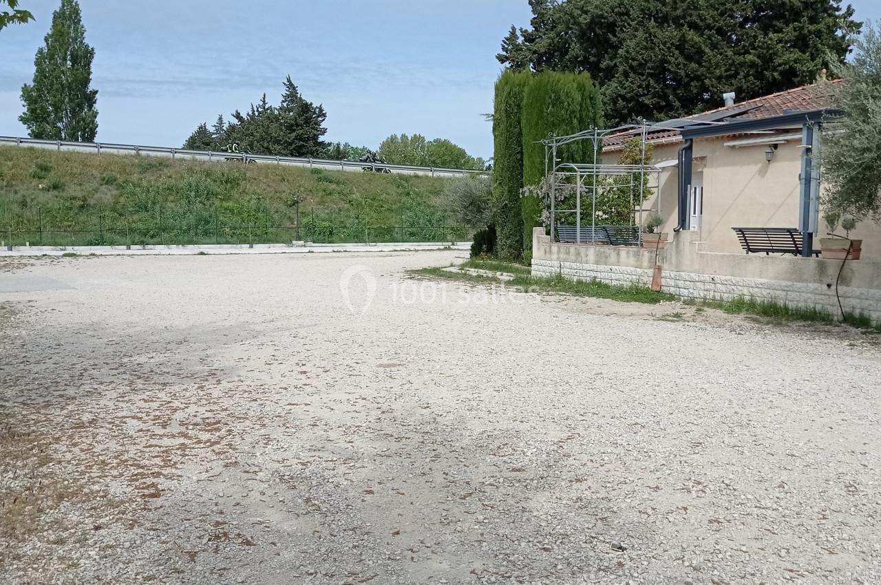 Cour en gravier bordée d'un bâtiment avec des arbres et une haie en arrière-plan, sous un ciel dégagé.