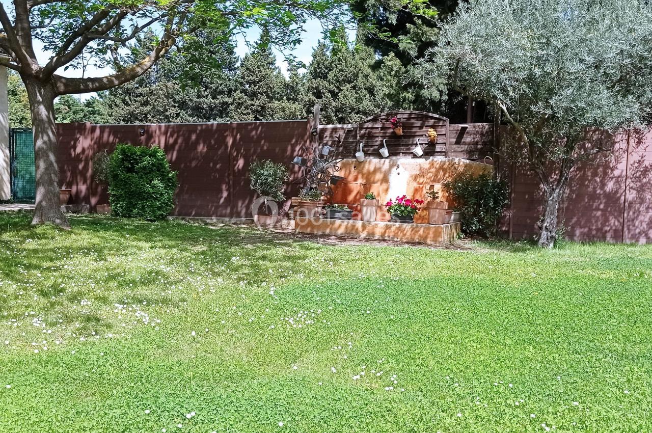 Jardin avec pelouse, fontaine en pierre ornée de pots de fleurs, arbres et mur en arrière-plan.