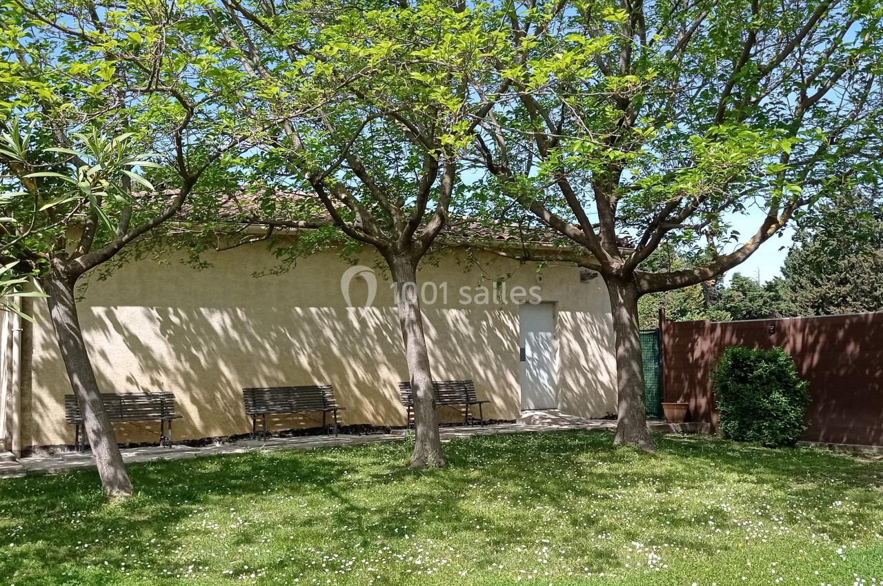 Maison beige entourée d'arbres avec des bancs à l'ombre, pelouse verte et clôture en bois à droite.
