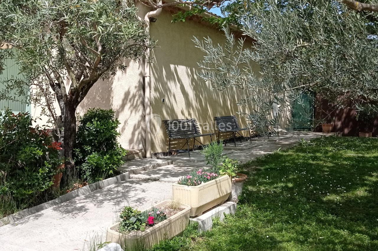 Jardin avec oliviers, pelouse, jardinières fleuries et bancs en bois adossés à une maison beige sous un ciel ensoleillé.