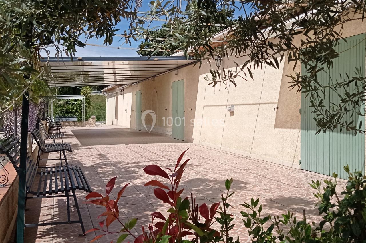 Terrasse dallée ombragée avec bancs, plantes et volets verts sur une façade beige sous un ciel dégagé.