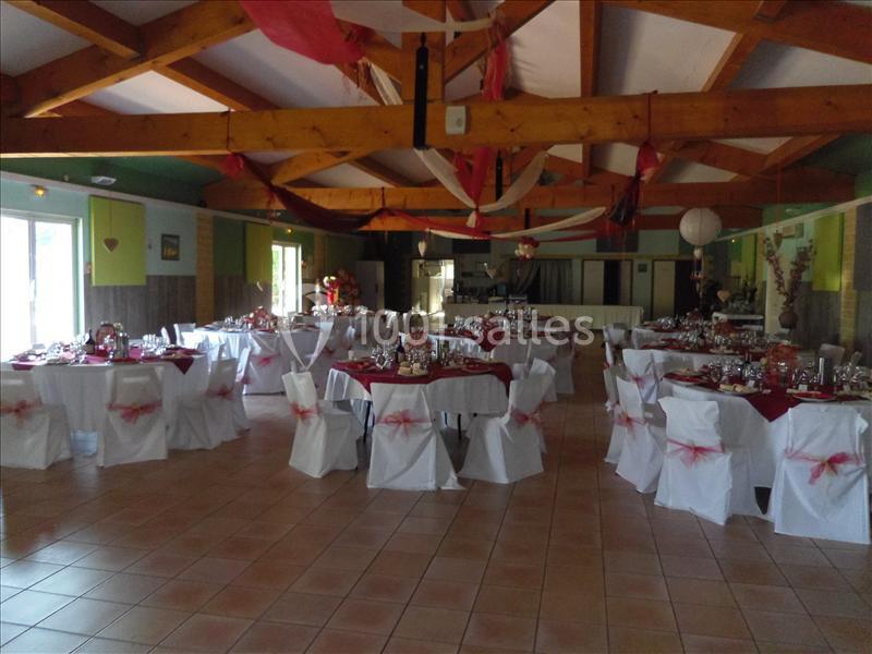 Salle décorée pour un événement avec tables dressées, nappes blanches et rubans rouges, sous un plafond en poutres…
