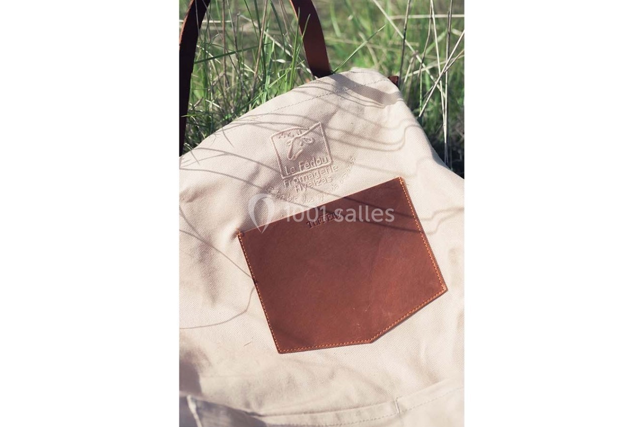 Sac en toile beige avec une poche en cuir marron et un logo embossé, posé sur de l'herbe.