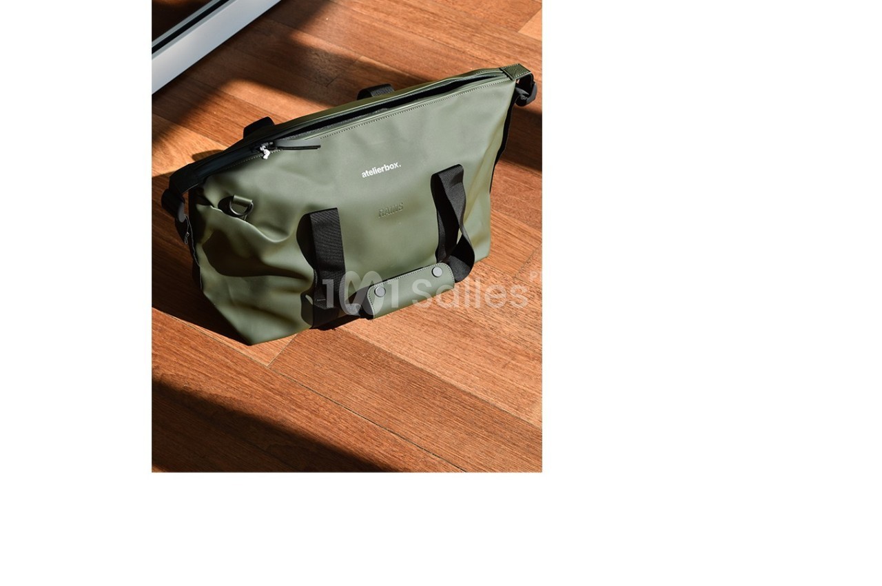Sac vert olive posé sur un sol en bois, avec des poignées noires et une fermeture éclair visible.