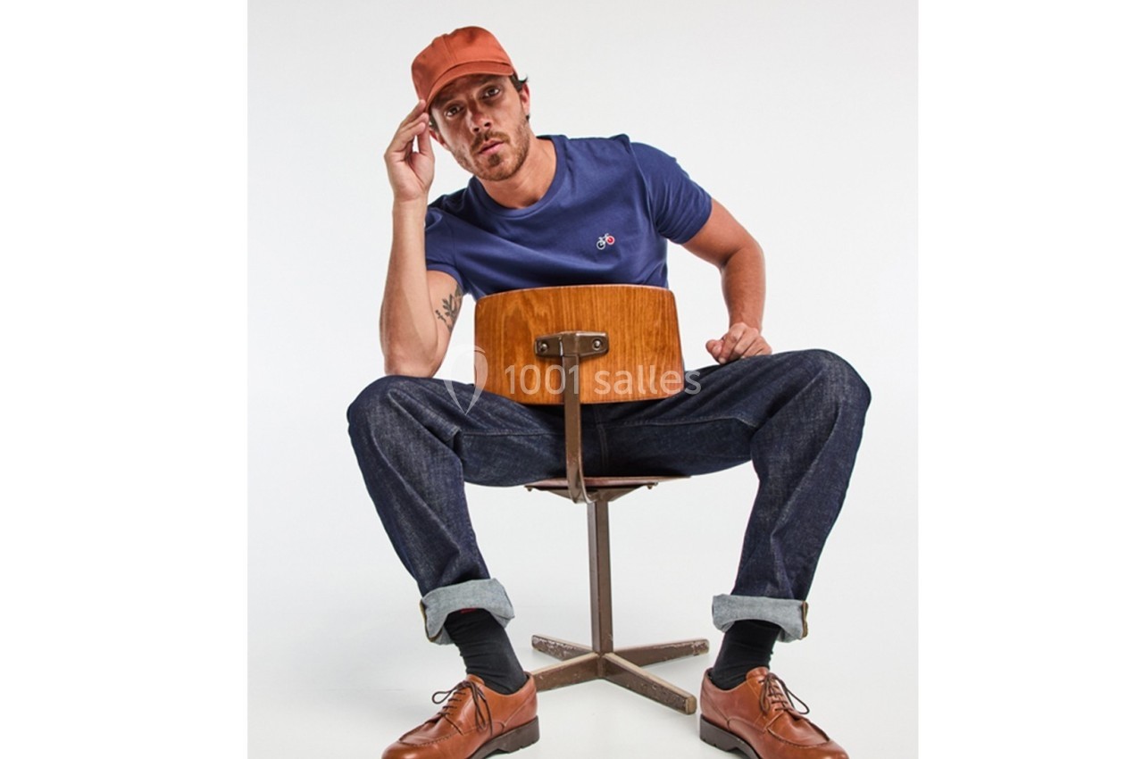 Homme assis à l'envers sur une chaise en bois, portant un t-shirt bleu, un jean, une casquette orange et des chaussures…