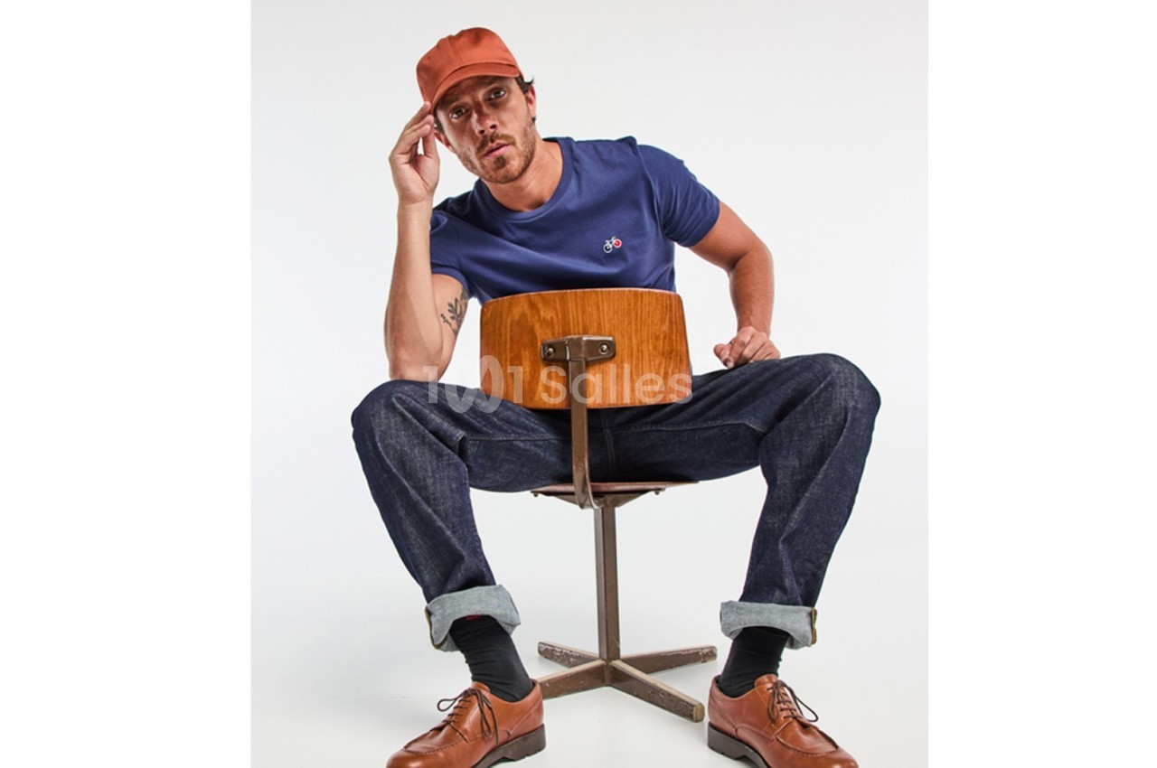 Homme assis à l'envers sur une chaise en bois, portant un t-shirt bleu, un jean, une casquette orange et des chaussures…