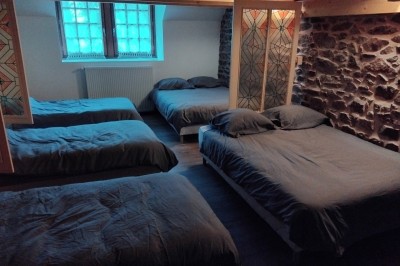 Bâtiment en pierre avec toit en ardoise, petite terrasse en bois, tables blanches et arbres en arrière-plan.