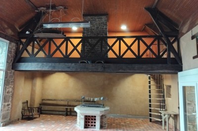 Bâtiment en pierre avec toit en ardoise, petite terrasse en bois, tables blanches et arbres en arrière-plan.