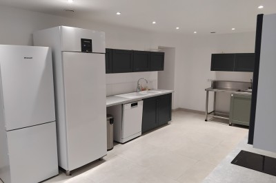 Bâtiment en pierre avec toit en ardoise, petite terrasse en bois, tables blanches et arbres en arrière-plan.
