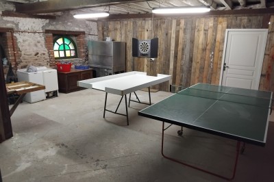 Bâtiment en pierre avec toit en ardoise, petite terrasse en bois, tables blanches et arbres en arrière-plan.