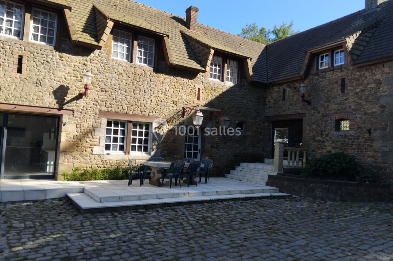 Cour pavée avec terrasse aménagée devant une maison en pierre à plusieurs fenêtres et toit en ardoise.