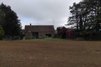 Bâtiment en pierre avec toit en ardoise, petite terrasse en bois, tables blanches et arbres en arrière-plan.