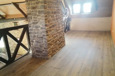 Bâtiment en pierre avec toit en ardoise, petite terrasse en bois, tables blanches et arbres en arrière-plan.