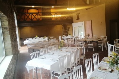 Bâtiment en pierre avec toit en ardoise, petite terrasse en bois, tables blanches et arbres en arrière-plan.