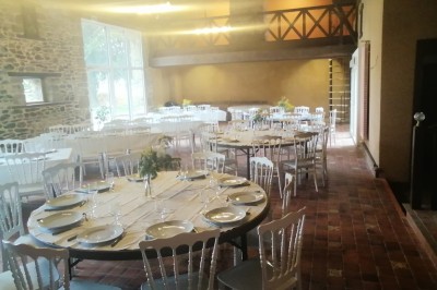 Bâtiment en pierre avec toit en ardoise, petite terrasse en bois, tables blanches et arbres en arrière-plan.