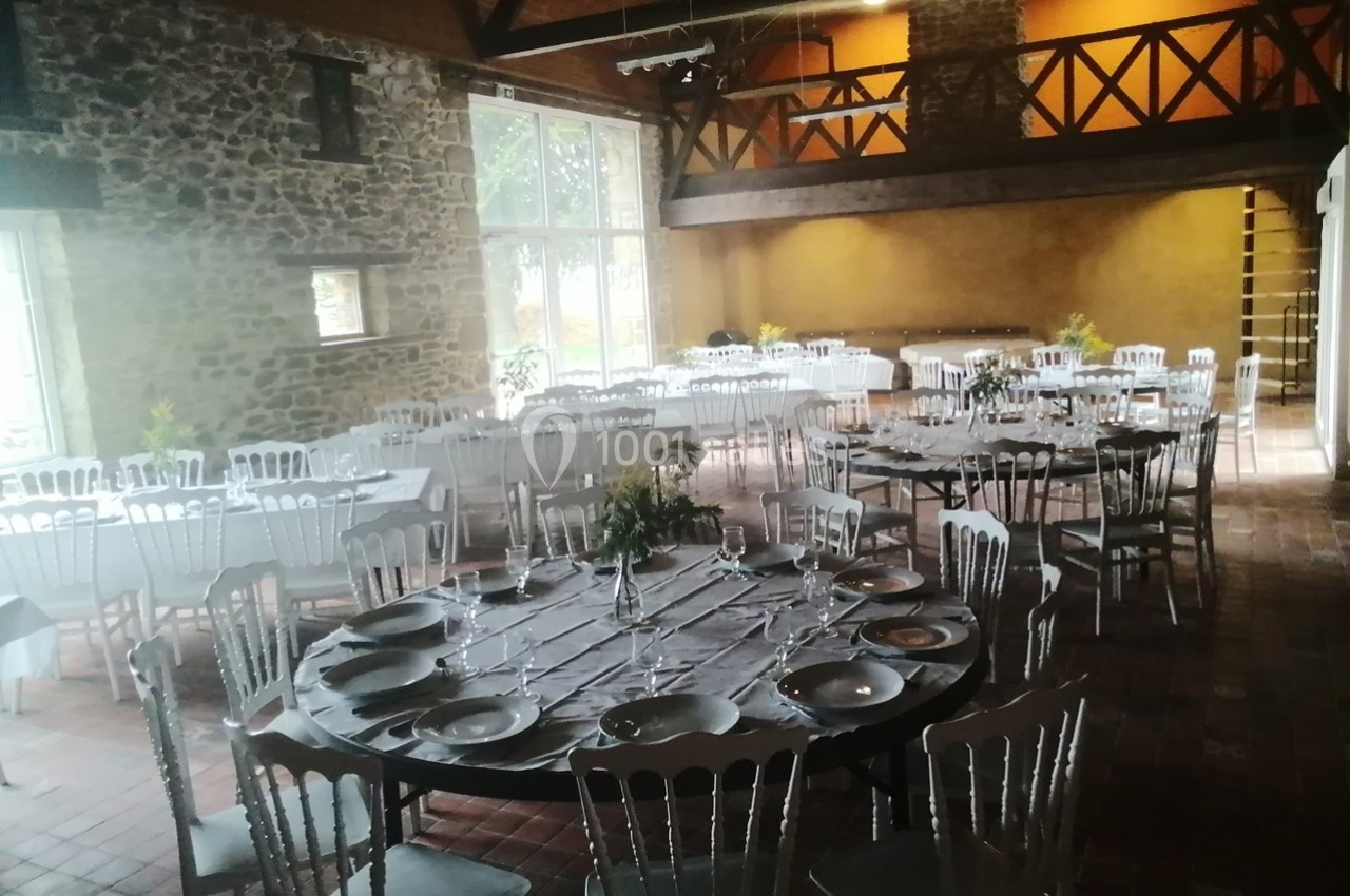 Salle de réception avec tables rondes et rectangulaires dressées, chaises blanches et murs en pierre apparente.