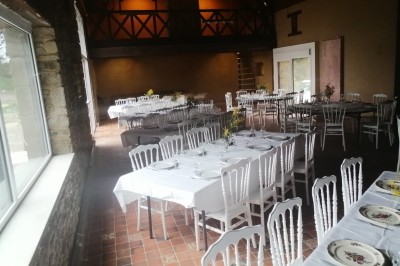 Bâtiment en pierre avec toit en ardoise, petite terrasse en bois, tables blanches et arbres en arrière-plan.