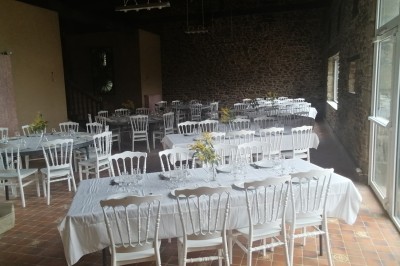 Bâtiment en pierre avec toit en ardoise, petite terrasse en bois, tables blanches et arbres en arrière-plan.
