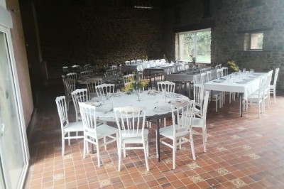 Bâtiment en pierre avec toit en ardoise, petite terrasse en bois, tables blanches et arbres en arrière-plan.