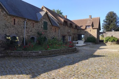 Bâtiment en pierre avec toit en ardoise, petite terrasse en bois, tables blanches et arbres en arrière-plan.